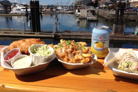A wooden table holds a plate of fish and chips with coleslaw and sauce, a loaded fries dish, a wrap sandwich, and a can of San Pellegrino. The table overlooks a marina with boats and a skyline in the background under a clear sky.