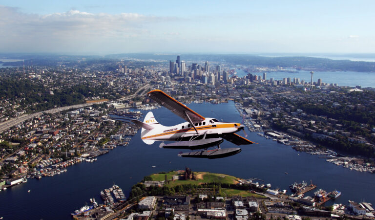 A seaplane flies over the bustling city of Seattle, with its soaring skyscrapers and expansive lake below. The skyline is dotted with tall buildings while green spaces and winding roads lay beneath a clear sky, offering a bird's-eye view of exciting things to do in this vibrant metropolis.