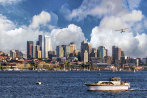 A city skyline with numerous modern skyscrapers under a partly cloudy sky. A small plane flies above the water, where several boats are visible, including a yacht in the foreground.
