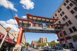A large traditional Chinese archway marks the entrance to the bustling Chinatown-International District, where city streets are lined with buildings, cars parked along the road, and a bright blue sky with scattered clouds overhead.