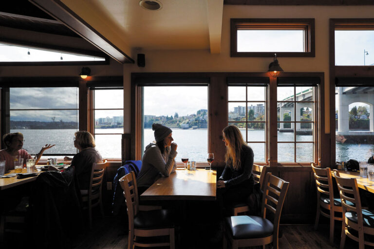 People sit and talk at wooden tables by large windows in a waterfront restaurant, enjoying a scenic view of the river, city buildings, and bridge connecting Bainbridge Island and Bremerton. Natural light brightens the cozy interior.