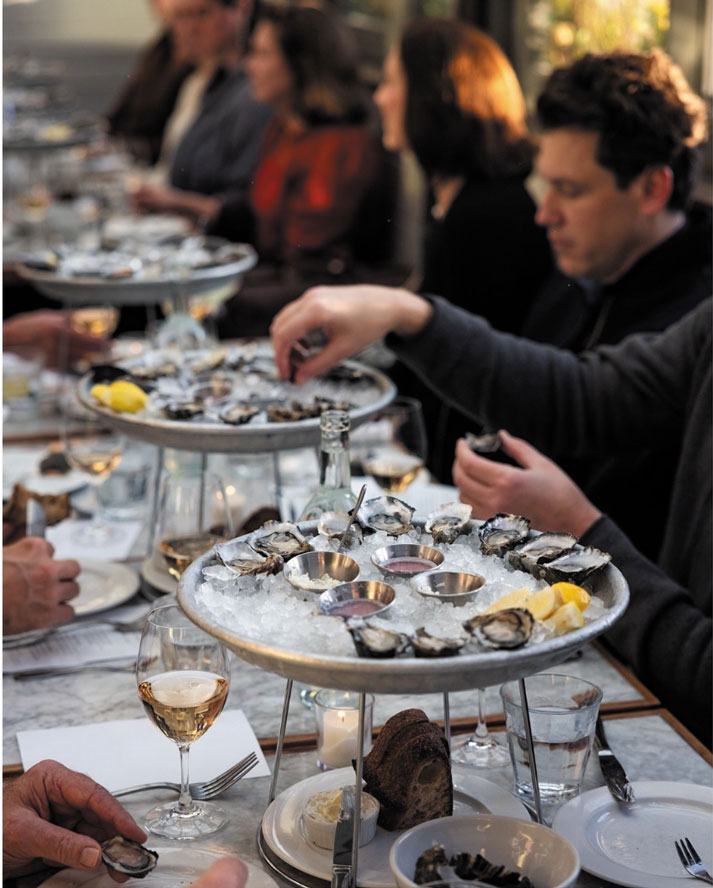 A group of people sit around a long table in the Ballard neighborhood, eating oysters from large platters on ice, with lemon wedges and sauces. Glasses of white wine and plates of bread add to the lively, social atmosphere.