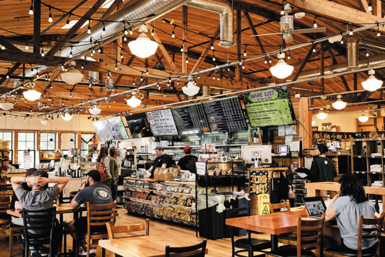 A bustling café with a wooden ceiling, hanging lights, and a central counter displaying menus and snacks, nestled in the lively Columbia City Neighborhood Seattle. Customers order at the counter as staff prepare food and drinks in a welcoming atmosphere.