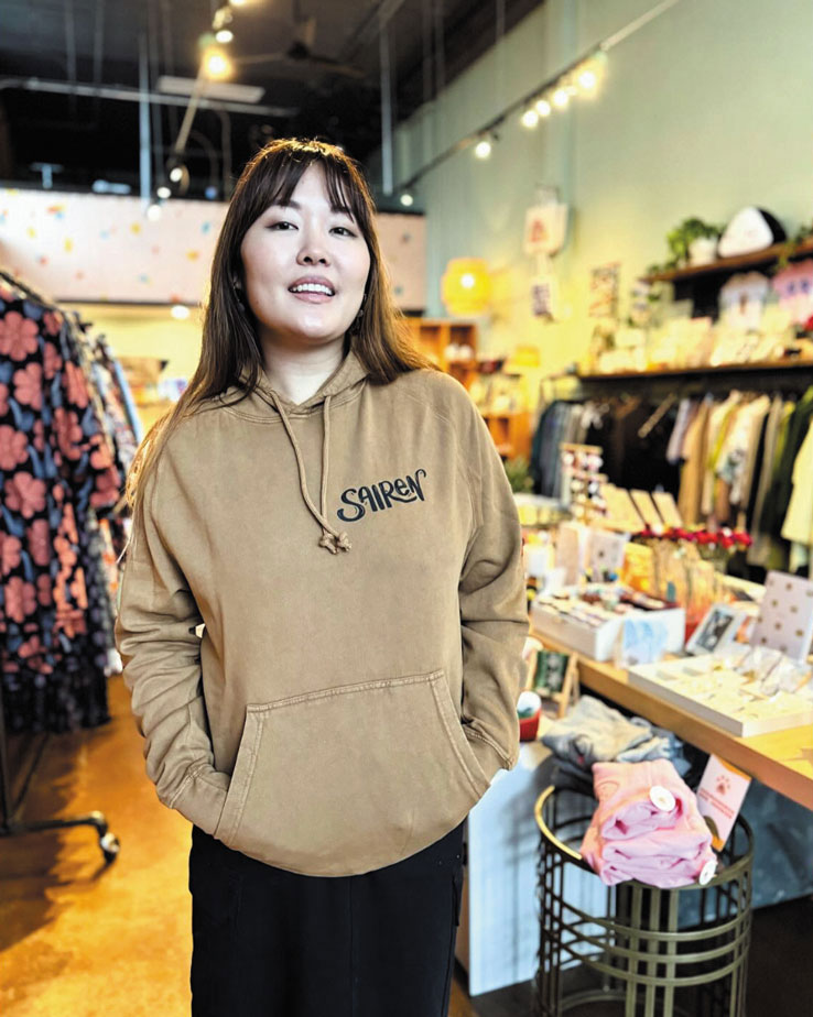 A woman wearing a tan “SIREN” hoodie stands smiling in a colorful Chinatown-International District boutique filled with clothes, gifts, and decor items displayed on racks and tables.