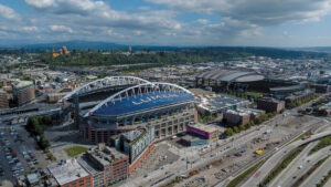 Aerial view of Lumen Field, a large stadium with a curved roof, nestled amid city buildings, roads, and greenery in Seattle’s SoDo neighborhood under a partly cloudy sky.