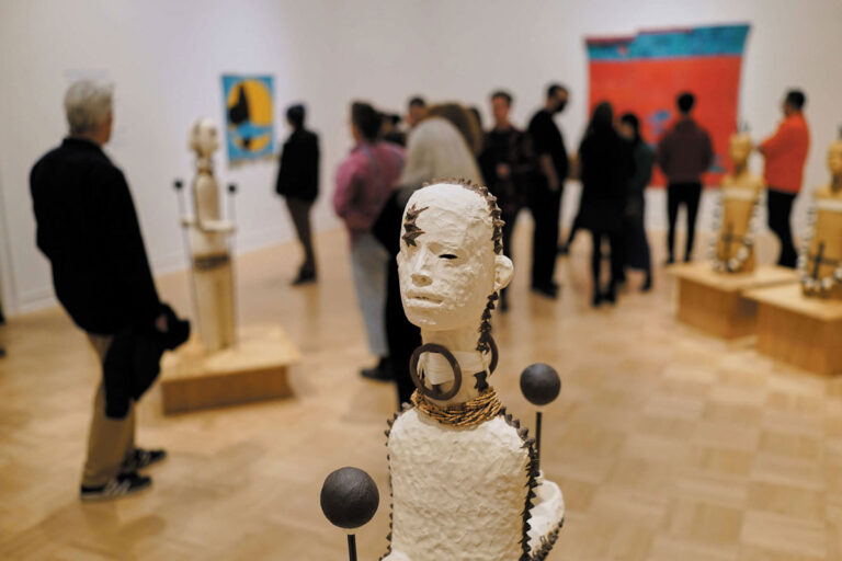 A group of people observe abstract sculptures and colorful paintings in an art gallery in the University District Seattle neighborhood. The foreground features a close-up of a textured humanoid sculpture with geometric details.