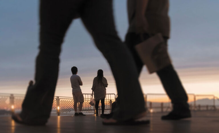 Two people stand near a railing at sunset in a waterfront neighborhood in Seattle, looking at the sky, while two blurred figures walk past in the foreground. The image has warm, soft lighting and a contemplative mood.