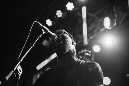 A black-and-white photo of a man passionately singing into a microphone on stage, surrounded by bright stage lights and a blurred background. He's wearing glasses and a dark t-shirt.