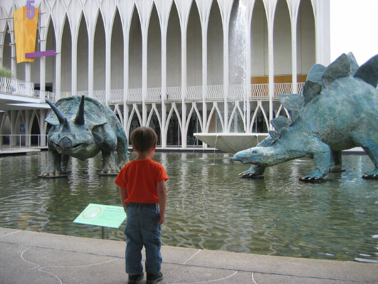 A child in an orange shirt stands in front of a pool with two large dinosaur sculptures, a Triceratops and a Stegosaurus, in an outdoor area with a building featuring tall arches in the background.