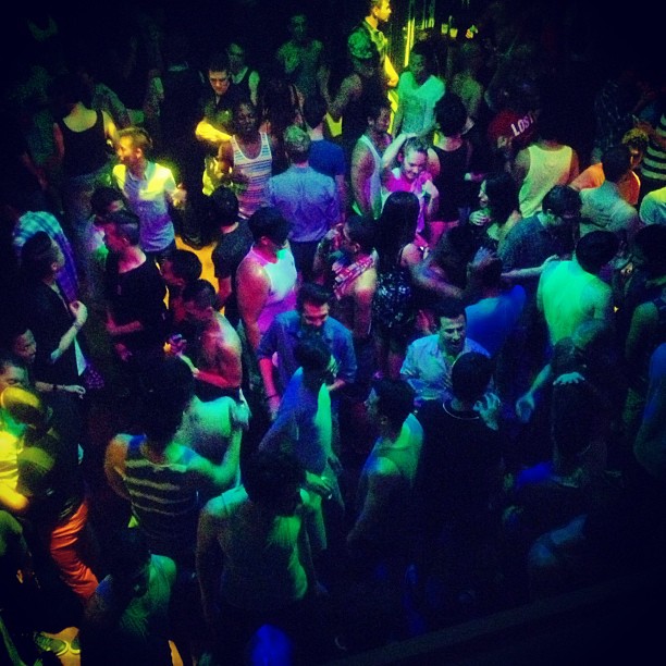 A vibrant, crowded nightclub scene with people dancing under colorful lights. The atmosphere is lively and energetic, with various individuals visible, enjoying the music and mingling in the dimly lit environment.