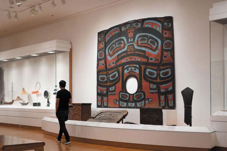 A person stands in a museum gallery in the downtown neighborhood of Seattle, viewing a large, colorful Indigenous artwork with bold patterns and a circular opening at its center, surrounded by display cases containing various artifacts.
