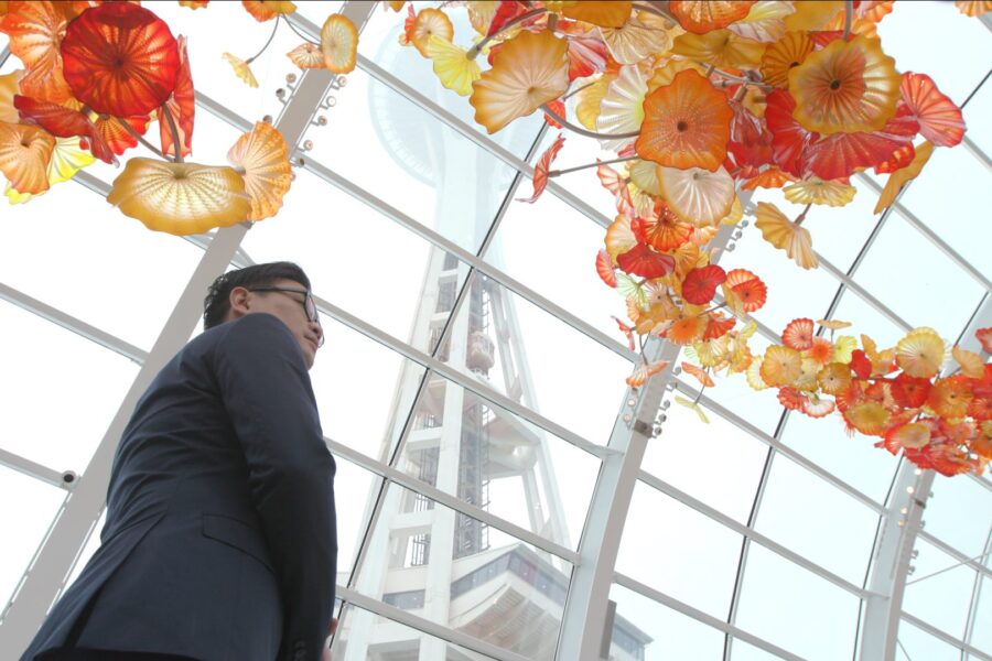 A person in a suit stands inside a glass building, looking up at colorful orange and yellow glass flower sculptures hanging from the ceiling. A tall structure is visible through the glass in the background.