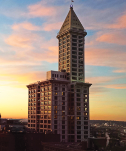 A tall, historic skyscraper with a distinctive pyramid-shaped top, set against a colorful sunset sky. The building is illuminated, reflecting warm tones, and surrounded by an urban cityscape.