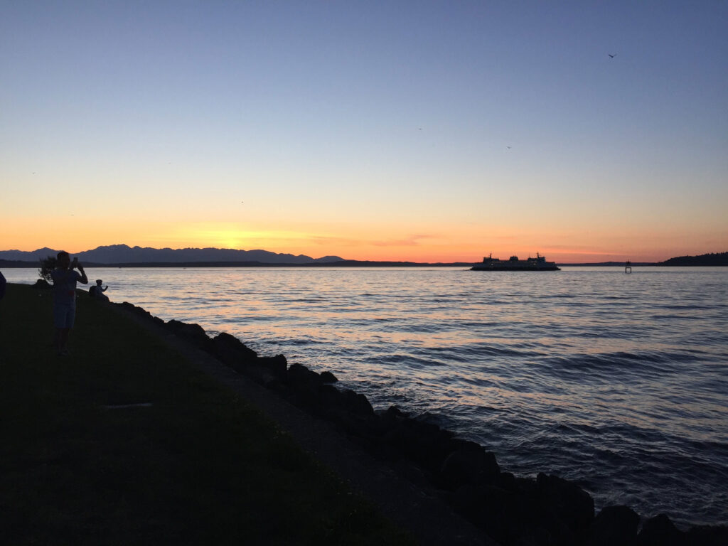 A serene sunset over a calm body of water, with a ferry in the distance. The sky is a gradient of orange and blue, and the shoreline is lined with rocks. A person stands on the left, capturing the view.