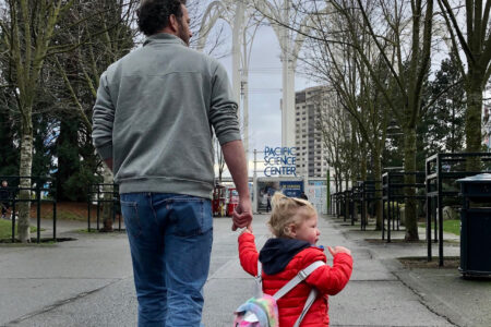 A man and a toddler walk hand in hand towards the Pacific Science Center. The toddler, wearing a red jacket and backpack with a unicorn design, points ahead. Trees line the path, and overcast skies loom above.