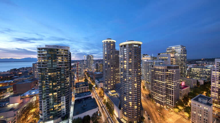 Aerial view of a cityscape at dusk reveals a mix of hotels for meetings of all kinds, with illuminated high-rise buildings overlooking bustling streets and a distant body of water under a clear blue sky turning to night.