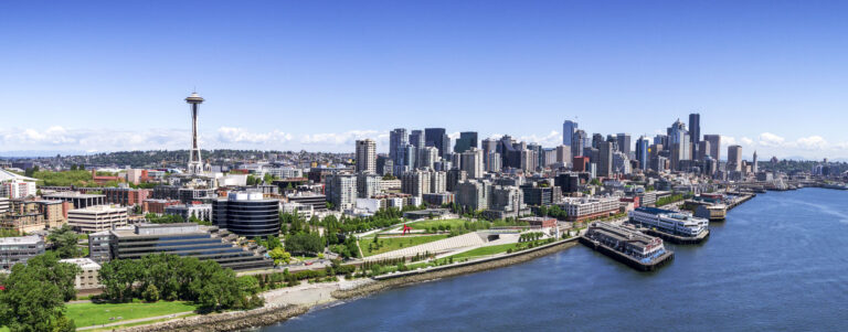 Aerial image of the Seattle waterfront featuring the green park area of the Olympic Sculpture Park in the foreground, the Space Needle to the left, and the continuing downtown skyline to the right, all under clear blue skies.