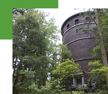 A round brick tower surrounded by lush green trees and shrubs. People are visible near the base of the tower, adding a sense of scale. The scene is serene with natural greenery.