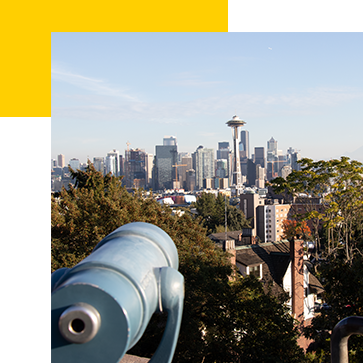 View of the Seattle skyline with the Space Needle prominently visible. A blue coin-operated telescope is in the foreground, and trees frame the urban landscape under a clear blue sky.