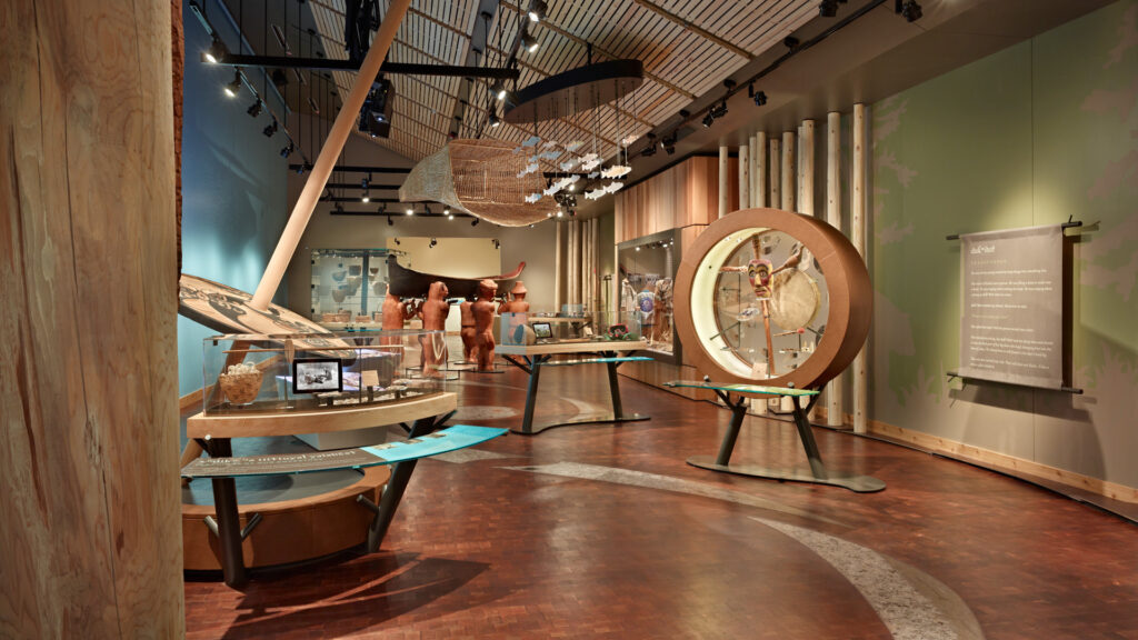 A modern museum exhibit with wooden accents displays artifacts, informational panels, and circular installations. The room features high ceilings and various indigenous art pieces, highlighted by soft lighting.