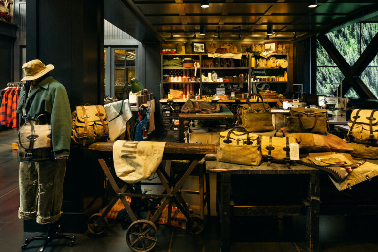 A rustic store interior featuring vintage-style bags, jackets, and hats displayed on wooden tables and racks. Dark wood and industrial elements create a cozy, nostalgic ambiance. Shelves at the back hold various leather goods and accessories.