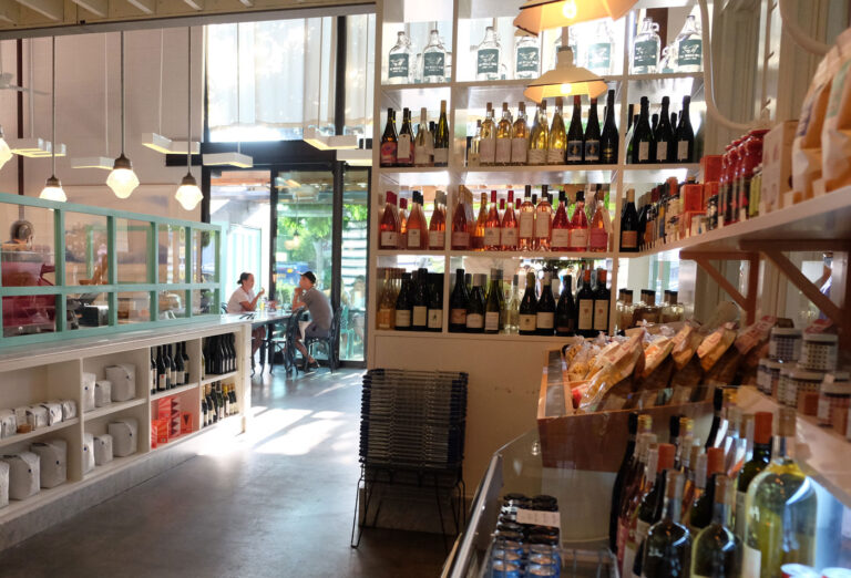 A cozy cafe interior with shelves displaying various bottles of wine and condiments. In the background, two people sit at a table near large windows letting in natural light. Stylish hanging lights add to the ambiance.