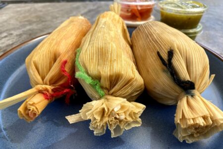 Three steamed tamales aligned on a plate with jars of condiments in the background.
