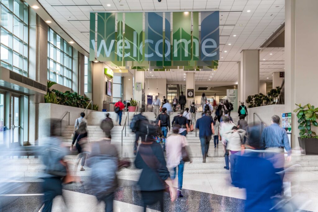 A bustling indoor scene shows people walking up a flight of stairs toward a large sign that reads "welcome." The area is bright and open, with plants and natural light from large windows. The setting suggests a busy public space or transportation hub.