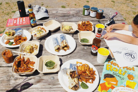 a spread of plates and takeout boxers with colorful food on a picnic table on the beach