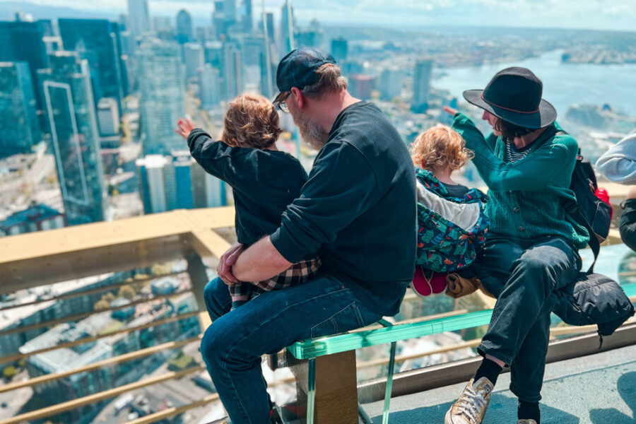 Two adults and two children sitting on a glass bench looking through large glass windows at the Seattle skyline.