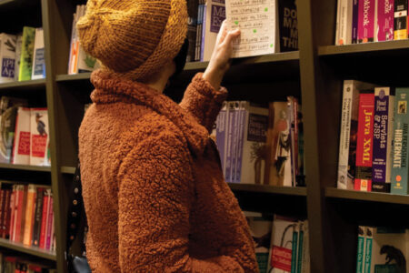 A person wearing an orange jacket and yellow beanie pulls out a book with a white cover from a bookshelf filled with books.