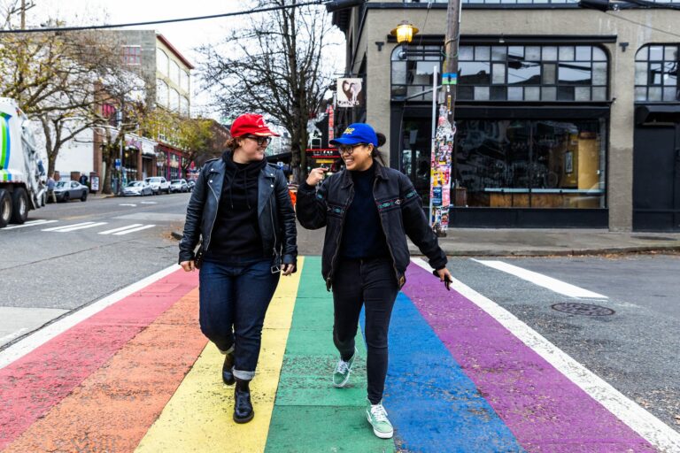 Two people walk across a vibrant rainbow crosswalk in a city. They are wearing casual clothing and hats, and are smiling at each other. Buildings and parked cars are visible in the background. A tree with bare branches is also in view.
