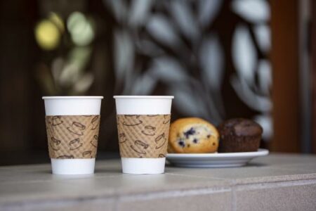 Two disposable coffee cups with brown sleeves are placed on a ledge. Behind them, a white plate holds a blueberry muffin and a chocolate muffin. The background is softly blurred with decorative leaf designs.