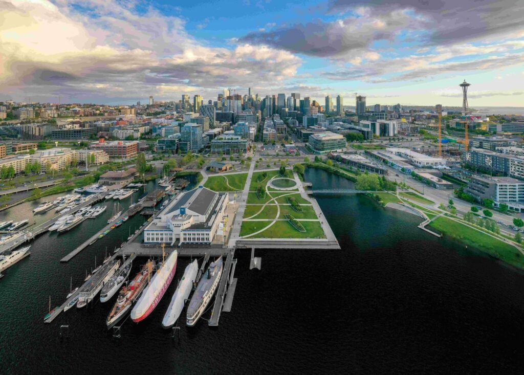 Aerial view of a cityscape featuring a waterfront with several docked ships. Numerous buildings and green spaces stretch toward a skyline with a distinctive tower. Clouds fill the sky above, and pathways crisscross the park area.