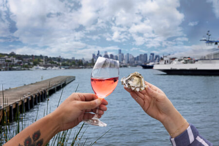 Two hands hold a glass of rosé wine and an oyster, with a scenic view of a city skyline and water in the background. A dock and ferry are visible under a partly cloudy sky.