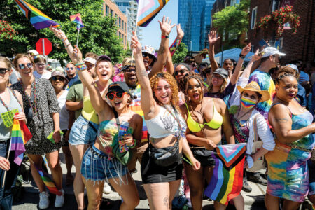 A vibrant crowd celebrates at a pride parade. People of various backgrounds are joyfully dressed in colorful outfits and waving rainbow flags, with smiles and arms raised in excitement under a sunny sky. Urban buildings and trees are visible.