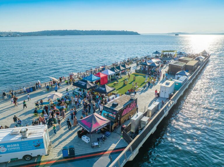 Aerial view of a bustling pier with food trucks, colorful tents, and people gathering. The pier extends over calm blue water under a clear sky. Activities and socializing create a lively atmosphere on this sunny day.