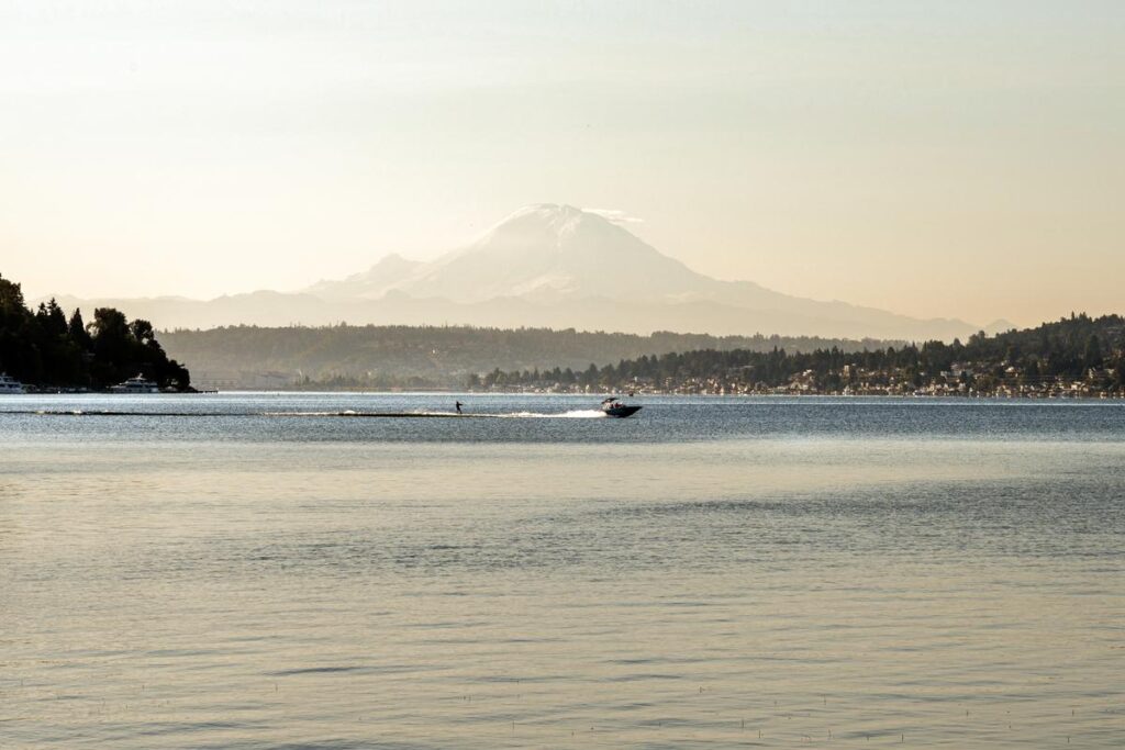 A serene lake with a person water skiing behind a boat. The horizon is lined with distant trees and Mount Rainier looms in the background under a clear sky. The atmosphere is calm and peaceful.