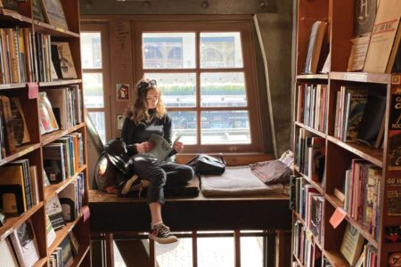 A person sits reading on a cozy window seat in a rustic bookstoresurrounded by shelves filled with books. Natural light streams in from the windowilluminating the relaxed scene.