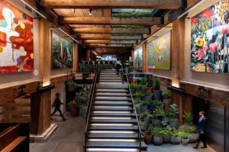 A modern interior with a wide staircase flanked by potted plants and large colorful abstract paintings on wooden walls, with people walking through the space.