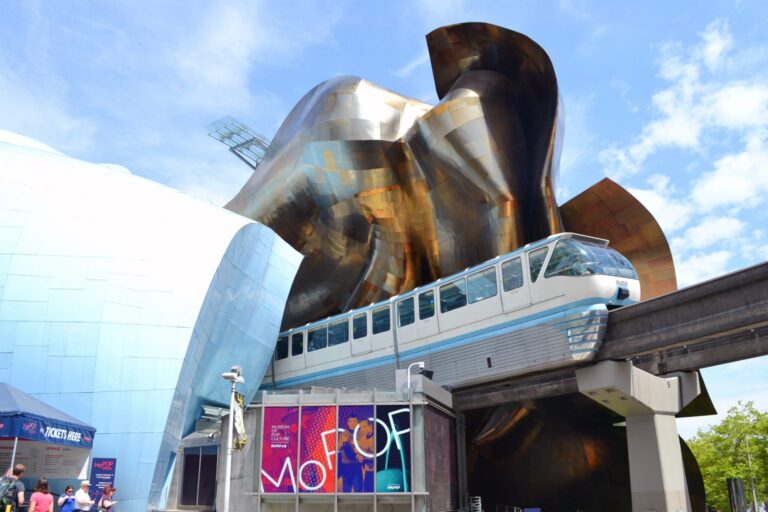 A monorail passes through the metallic, curved exterior of the Museum of Pop Culture in Seattle on a sunny day, with a blue sky and people gathered near the entrance.