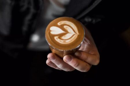 A person holding a cup of coffee topped with intricate latte art featuring a heart and leaf pattern, viewed from above. The person wears a dark jacket and white shirt.