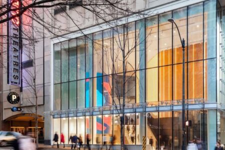 A city street scene with people walking past a modern building with large glass windows. Inside, colorful abstract artwork is visible. There is a tree with bare branches and a “THEATRES” sign on the left side.