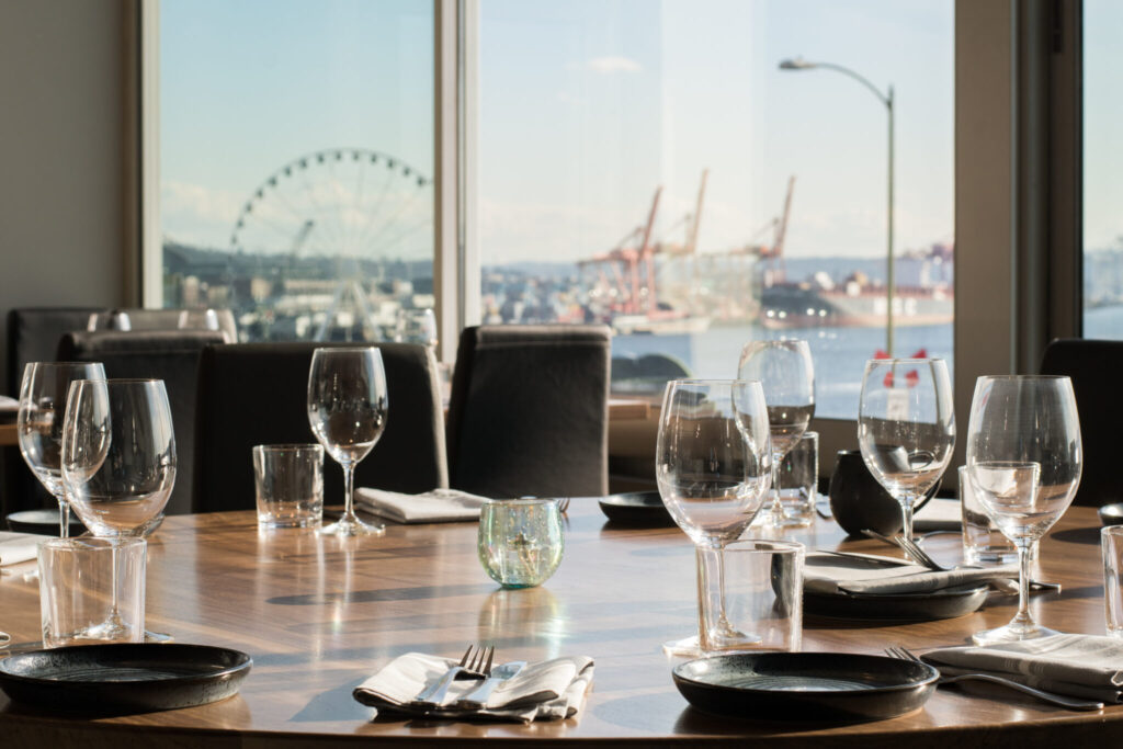 A set dining table with empty wine glasses and plates in a restaurant, large windows showing a view of a ferris wheel, water, and industrial cranes outside.