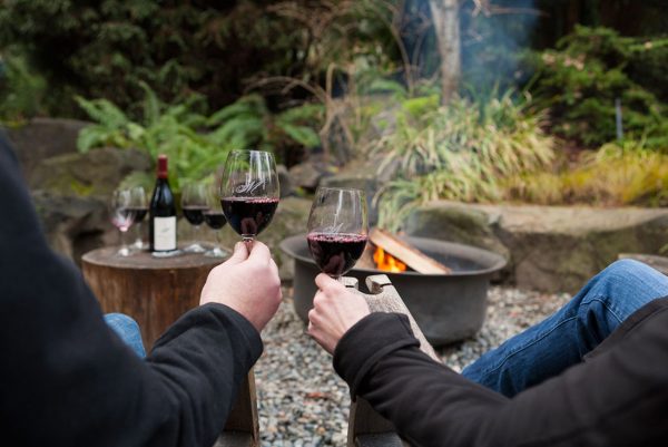 Two people relax by a fire pit outdoors, holding glasses of red wine. A bottle of wine and another glass sit on a wooden stump nearby, surrounded by greenery and rocks.