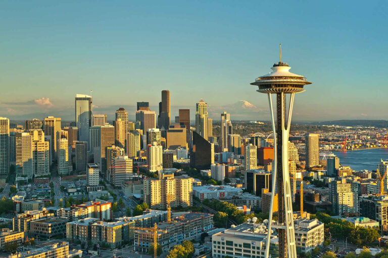 Seattle skyline at sunset featuring the Space Needle in the foreground, modern skyscrapers, and Mount Rainier visible in the background under a clear sky.
