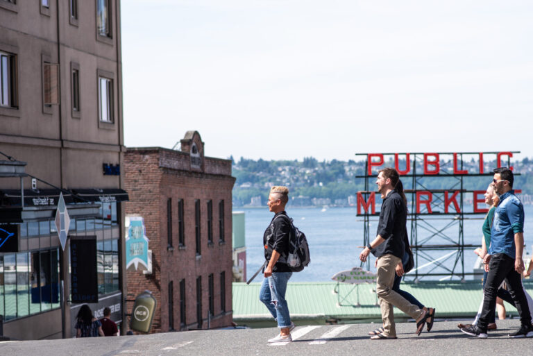 Four people cross a city street with a view of water and distant trees. A "Public Market" sign is visible in the background, hinting at a popular marketplace near the waterfront. It is a bright, sunny day.