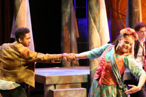 Two young actors perform on stage in colorful costumes, holding hands as they dance. The background features columns and dramatic lighting, suggesting a theatrical or historical setting.