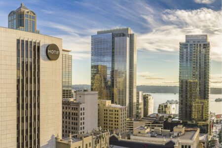 Downtown cityscape featuring tall modern buildings, including one with a “MOTIF” sign, under a partly cloudy sky with water and distant land visible in the background.