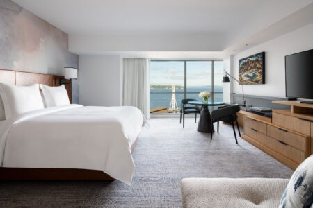 A modern hotel room with a large bed, desk, TV, and dresser. Floor-to-ceiling windows reveal a view of the waterfront, ferris wheel, and blue sky. The décor is neutral with wood accents and a patterned carpet.